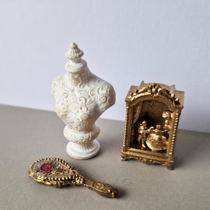 Puede incluir: Un conjunto en miniatura que incluye un busto de color crema con volutas ornamentadas, un marco rectangular dorado con un relieve de dos figuras y un pequeño espejo de mano dorado decorado con un motivo de rosa roja y hojas verdes.
