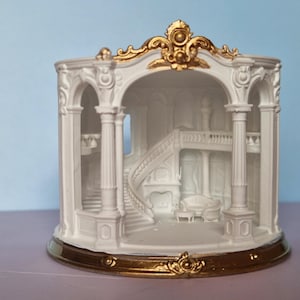 Könnte beinhalten: Ein weißes Keramik-Dekorationsobjekt mit einer detaillierten Innenszene, die eine geschwungene Treppe, Säulen und Möbel zeigt. Die Struktur ist mit einem goldenen, verzierten Rand versehen und steht auf einem passenden goldenen Sockel.