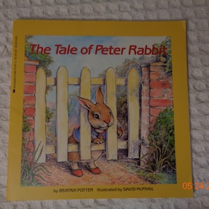 May include: A yellow book cover with the title "The Tale of Peter Rabbit" in red text. The cover features an illustration of a brown rabbit peeking through a white picket fence. The book is by Beatrix Potter and illustrated by David McPhail.
