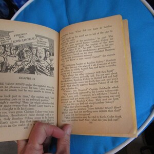 May include: A vintage book open to a page with a black and white illustration of a group of people holding signs that say "Give the Bonds with Earth" and "Freedom for Alpha Centauri". The text on the page reads "Chapter 15" and "There were rings under the captain's eyes, there had been no pleasure jaunt for him. Larry saluted as crisply as he could, but he was unable to conceal his fatigue."