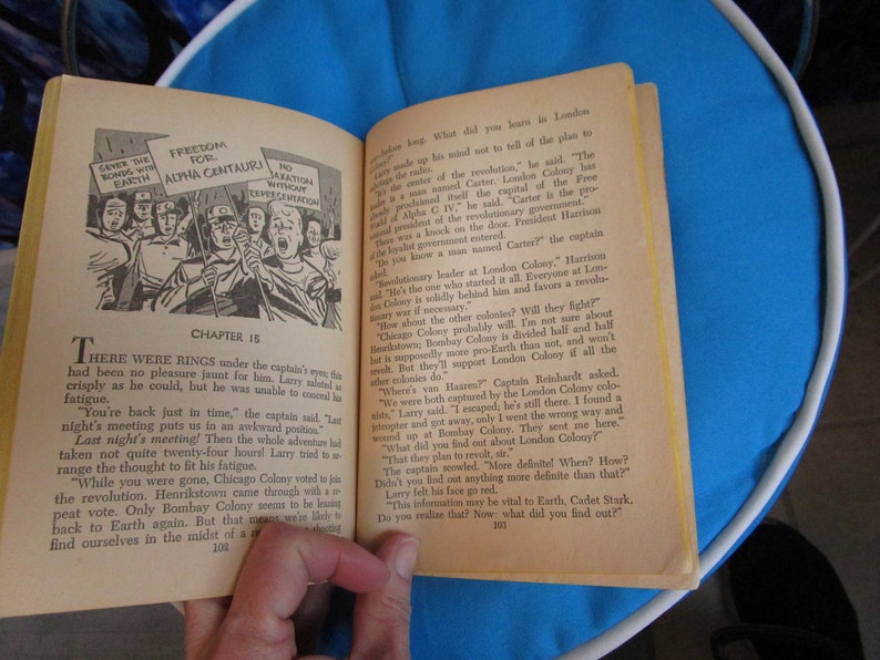 May include: A vintage book open to a page with a black and white illustration of a group of people holding signs that say "Give the Bonds with Earth" and "Freedom for Alpha Centauri". The text on the page reads "Chapter 15" and "There were rings under the captain's eyes, there had been no pleasure jaunt for him. Larry saluted as crisply as he could, but he was unable to conceal his fatigue."