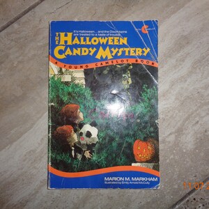 May include: A blue book cover with the title "The Halloween Candy Mystery" and the subtitle "A Young Camelot Book". The cover features an illustration of two children dressed in costumes looking at a figure in the bushes. The book is by Marion M. Markham and illustrated by Emily Arnold McCully.
