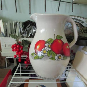 May include: A cream-colored ceramic pitcher with a handle and a spout. The pitcher is decorated with a design of three red apples, green leaves, and small white daisies. The pitcher is sitting on a white wire shelf.