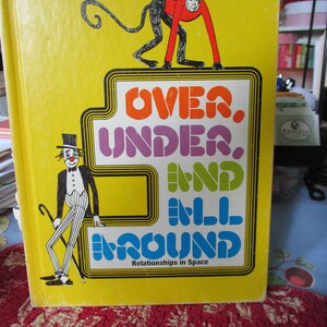 May include: A vintage yellow book titled "OVER, UNDER, AND ALL AROUND" with a cartoon monkey and clown illustration. The book's cover features the text "Relationships in Space" and is set against a red floral patterned background.
