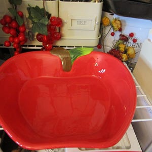 May include: A large, glossy red ceramic apple-shaped bowl with a brown stem and green leaf. The bowl is sitting on a white wire shelf. Decorative items are in the background.