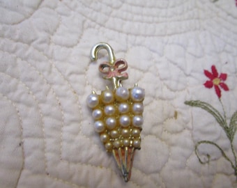 May include: A vintage umbrella brooch with a gold-tone metal frame. The umbrella's canopy is adorned with faux pearls, and a small pink bow accents the handle. The bottom of the umbrella has a painted, colorful design.