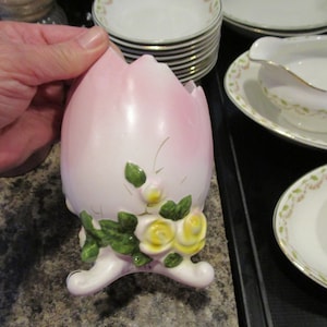 May include: A pink ceramic egg-shaped dish with three feet and floral designs. The dish has green leaves and yellow flowers.