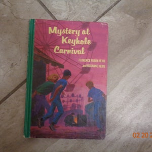 May include: A pink and green paperback book titled "Mystery at Keyhole Carnival" by Florence Parry Heide and Roxanne Heide. The cover features an illustration of three children walking through a carnival.