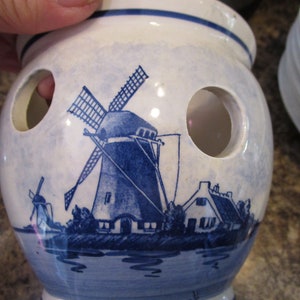 Puede incluir: Un portacebollas de cerámica blanca con un diseño de Delftware azul y blanco que presenta un molino de viento y casas. El portacebollas tiene varios agujeros para guardar cebollas.