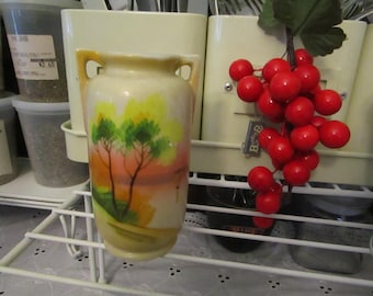May include: A white ceramic vase with a painted scene of trees and a sunset. The vase has a rounded body and two handles. The vase is sitting on a white shelf with a bunch of red berries hanging from a green plant.