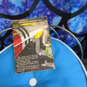 May include: A vintage paperback book with a black and white illustration of a rocket ship and an astronaut in space. The book's cover reads "SCHOLASTIC BOOK SERVICES, NEW YORK" and includes a description of the story on the back cover.