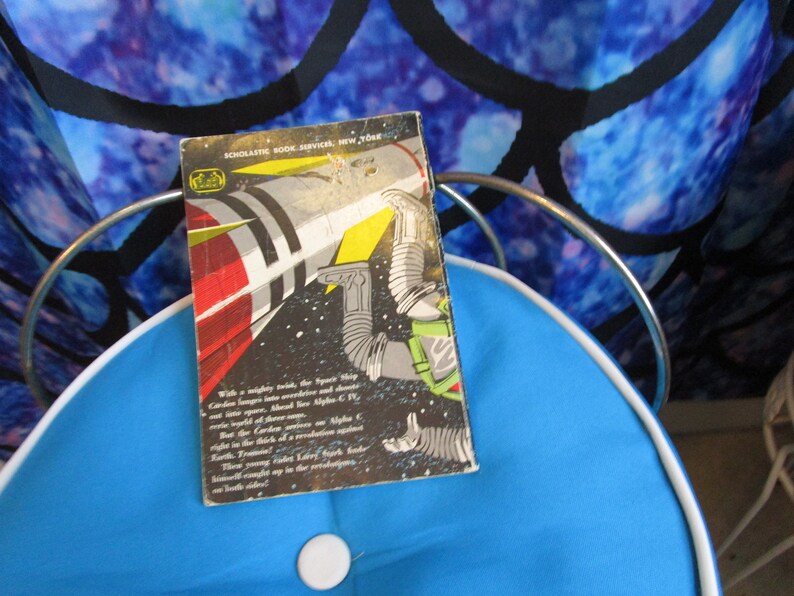 May include: A vintage paperback book with a black and white illustration of a rocket ship and an astronaut in space. The book's cover reads "SCHOLASTIC BOOK SERVICES, NEW YORK" and includes a description of the story on the back cover.