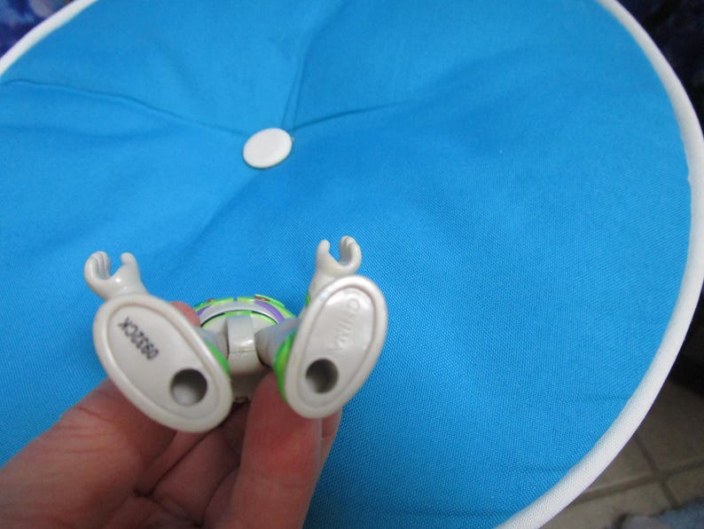 May include: A Buzz Lightyear action figure, lying on its back, with its feet pointing towards the viewer. The figure is primarily white, green, and purple, with the number "09320K" visible on one foot. The figure is resting on a blue cushion.