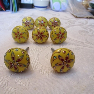 May include: A collection of eight yellow cabinet knobs with a floral design. Each knob features a glossy finish and is adorned with red, white, and green accents. The knobs are round with a metal stem for installation.