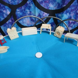 May include: Miniature dollhouse furniture set, including a dresser, bathtub, washing machine, table, and stool. The furniture is off-white, with a red and white stool. The items are arranged on a blue surface.