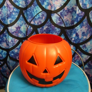 May include: Orange plastic pumpkin shaped Halloween trick-or-treat bucket with a black handle and a carved jack-o'-lantern face.