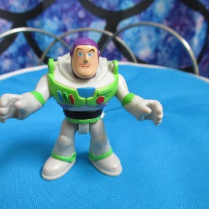 May include: A Buzz Lightyear action figure, featuring a white and green spacesuit with a purple helmet and a smiling face. The toy is standing on a blue surface, with a decorative background.