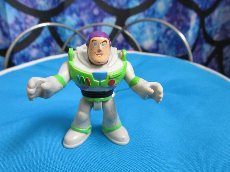 May include: A Buzz Lightyear action figure, featuring a white and green spacesuit with a purple helmet and a smiling face. The toy is standing on a blue surface, with a decorative background.