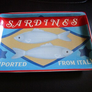 May include: A rectangular ceramic tray featuring a graphic design of two light blue fish centered on a tan diamond shape. The tray has a red border and blue background with the text SARDINES at the top and IMPORTED FROM ITALY at the bottom.