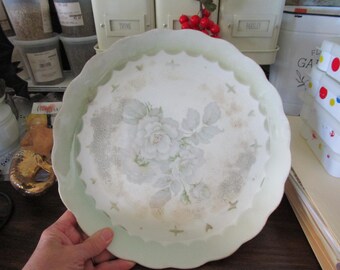 May include: A white porcelain serving dish with a scalloped edge and a green rim. The dish features a floral design with white roses and green leaves on a white background.