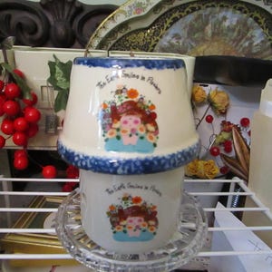May include: A ceramic candle warmer featuring a two-piece design with a cream-colored base and lid. Both pieces are decorated with a blue sponge-painted rim and a central graphic of a person surrounded by flowers with the text The Earth Smiles in Flowers.