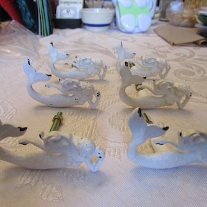 May include: Six white mermaid cabinet knobs with black accents. The mermaid figures are in a reclining pose with their tails curved. The knobs are made of a solid material and are designed for furniture or decorative use.
