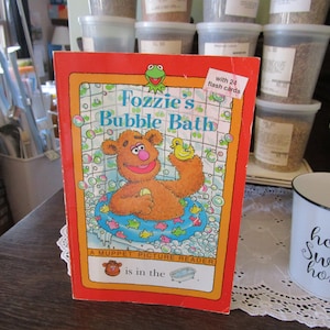 May include: A red and orange children's book titled "Fozzie's Bubble Bath" featuring a cartoon illustration of Fozzie Bear taking a bath. The book is a Muppet Picture Reader and includes 24 flash cards.