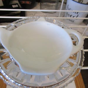May include: A white ceramic teapot-shaped tea bag holder. The holder sits on a clear, cut-glass saucer with a decorative pattern. The teapot has a spout and handle. The background includes a white shelf and other kitchen items.