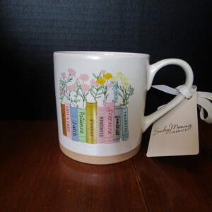 May include: A white ceramic mug featuring a colorful illustration of a row of books with flowers growing from the top. Each book spine is labeled with words including Faith, Patience, Prayer, Kindness, Strength, and Goodness. A tag reads Sunday Morning Ceramics.
