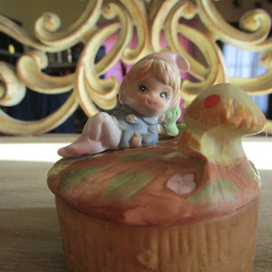 May include: A ceramic trinket box with a whimsical design featuring a small girl figurine sitting on a mushroom. The girl is wearing a blue dress and a pink hat. The mushroom is brown with a red spot on top. The box is decorated with a brown, green, and pink floral pattern.