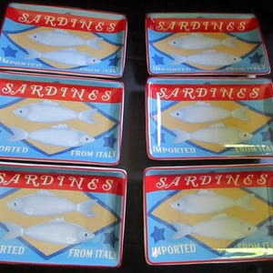 May include: A set of six rectangular ceramic plates featuring a graphic design of two white fish. Each plate displays the red text SARDINES at the top and the blue text IMPORTED FROM ITALY at the bottom, set against a light blue and yellow background.