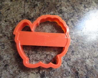 May include: An orange plastic turkey-shaped cookie cutter. The cutter has a rectangular handle with the word "TURKEY" printed on it. The brand name "Wilton" is also visible on the handle.