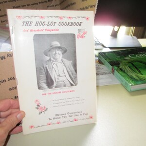 May include: A vintage cookbook titled "The Hog-Lot Cookbook" with a black and white portrait of a man wearing a hat. The cover has red floral accents and text that reads "And Household Companion". Another book with a green cover is visible.