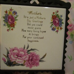 May include: A square ceramic tile with a floral design and the words "Mother" and a Mother's Day message. The tile is framed in a black, decorative metal frame. Pink and white flowers are at the bottom of the tile.