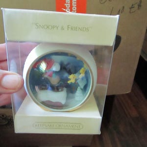 May include: A round Snoopy & Friends Keepsake Ornament encased in a clear plastic box. The ornament features a miniature scene with Snoopy wearing a red hat, sitting on a white snowy base with small yellow star figures against a dark blue background.