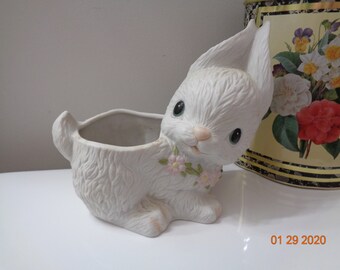 May include: White ceramic planter shaped like a rabbit with pink flowers around its neck. The planter has a square opening for planting.