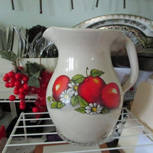 May include: Cream-colored ceramic pitcher with a handle, featuring a vibrant apple design with red apples, green leaves, and white daisies. The pitcher is a decorative kitchenware item.