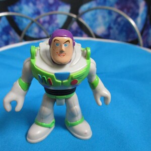 May include: A Buzz Lightyear action figure, featuring a white and green spacesuit with purple accents. The toy has a smiling face and is posed on a blue surface. The figure is a collectible toy.