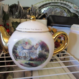 May include: White ceramic teapot with a gold handle and spout. The teapot features a scenic illustration of a house and the text "Home Is Where The Heart Is". The lid is topped with a gold knob.