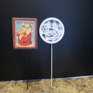 May include: A rectangular garden stake featuring a vintage-style Santa Claus illustration with the text Happy Holidays and a circular white garden stake featuring a cutout design of two birds perched on a branch against a checkered background.