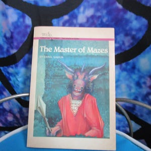 May include: A paperback book titled "The Master of Mazes" by Carol Daskin. The cover features a cartoon illustration of a horned creature in a red robe holding a spear. The background is a teal and green color.