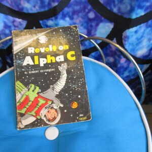 May include: A vintage paperback book titled "Revolt on Alpha C" by Robert Silverberg. The cover features a colorful illustration of an astronaut in space.