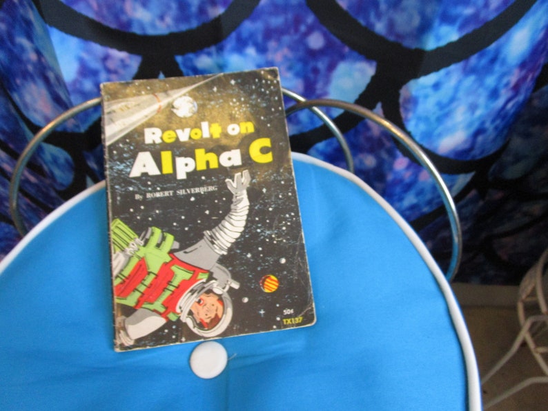 May include: A vintage paperback book titled "Revolt on Alpha C" by Robert Silverberg. The cover features a colorful illustration of an astronaut in space.
