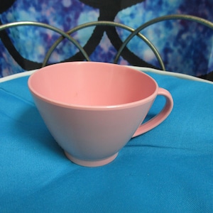 May include: A pink plastic teacup with a handle. The cup is sitting on a blue surface.