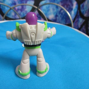May include: A Buzz Lightyear action figure, viewed from the back, stands on a blue surface. The toy features a white and green spacesuit with a purple head and black accents. The figure is a collectible item.