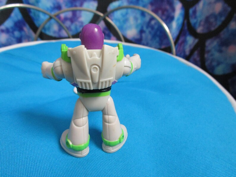 May include: A Buzz Lightyear action figure, viewed from the back, stands on a blue surface. The toy features a white and green spacesuit with a purple head and black accents. The figure is a collectible item.