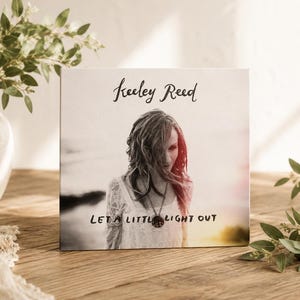 May include: A square canvas print featuring a black and white photograph of a person with long hair wearing a lace top and a pendant necklace. The text Keeley Reed appears at the top, and the text Let a little light out is at the bottom.