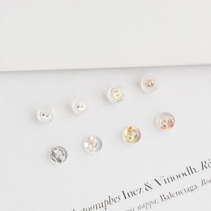 May include: A selection of clear, round earring backs with different metal finishes, including silver, gold, and rose gold. The earring backs are made of a transparent material, allowing the metal details to be seen. The image is taken from a high-angle perspective.
