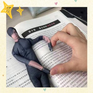 May include: A small, dark gray doll in a full body suit with a mesh face and hands is positioned on top of an open book. The book has Chinese text and is open to a page with diagrams and text.