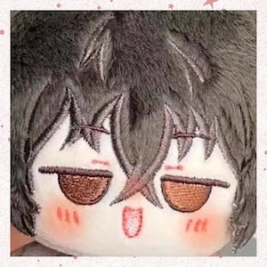May include: A plush doll with embroidered facial features. The doll has dark gray fur-like hair and a white face with brown eyes, rosy cheeks, and a small open mouth. The doll's expression appears sleepy or relaxed.